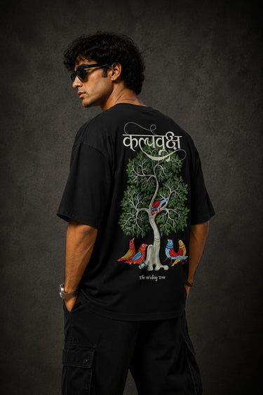 Man wearing a black Kalpvruksh oversized t-shirt with a colorful tree and bird graphic print on the back. Features Hindi script and 'The Wishing Tree' text. Streetwear style.