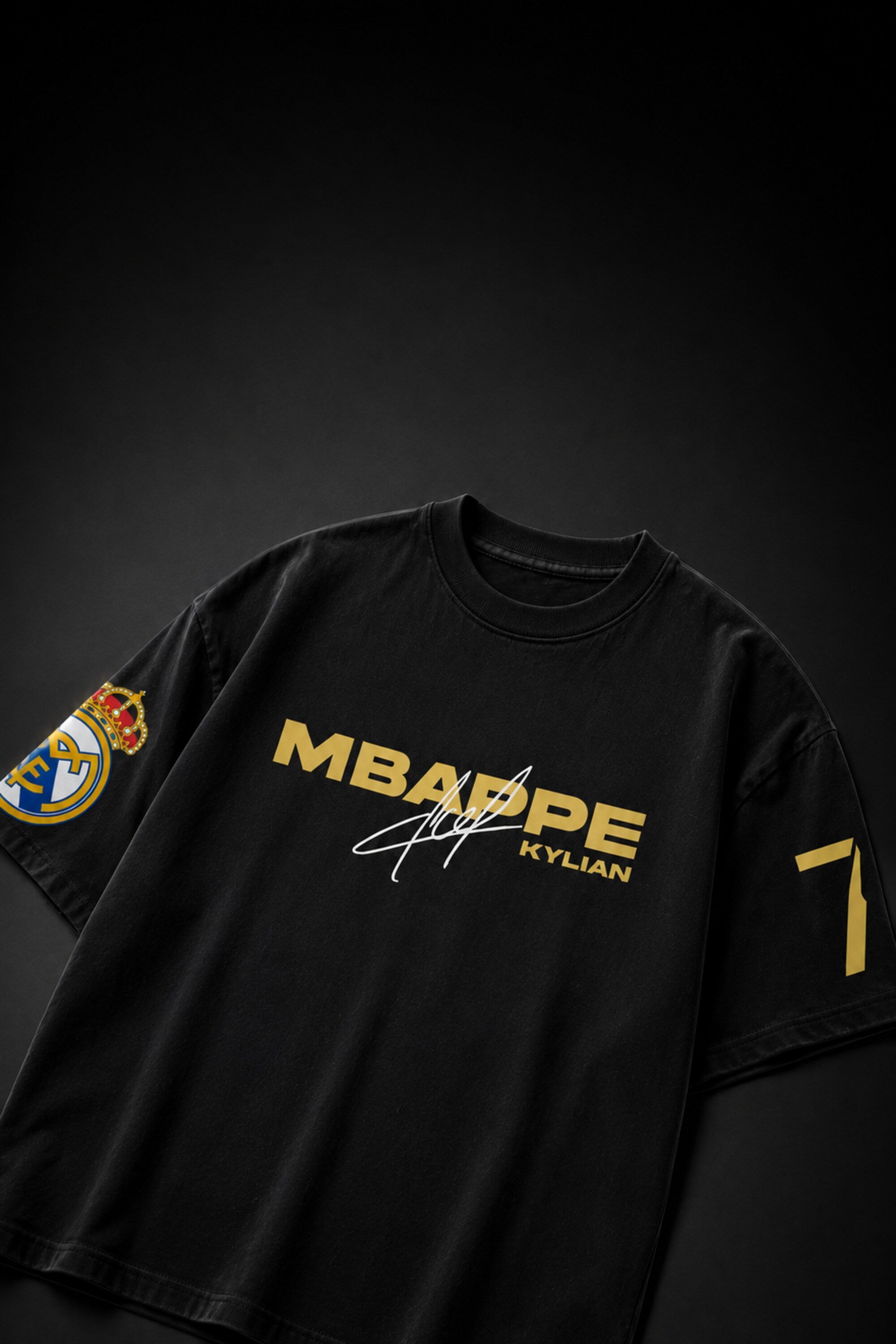 A black short-sleeve t-shirt featuring 'MBAPPE KYLIAN' printed in gold across the chest, with a white signature graphic below it. The left sleeve features a Real Madrid crest, and the right sleeve displays the number 7 in gold.