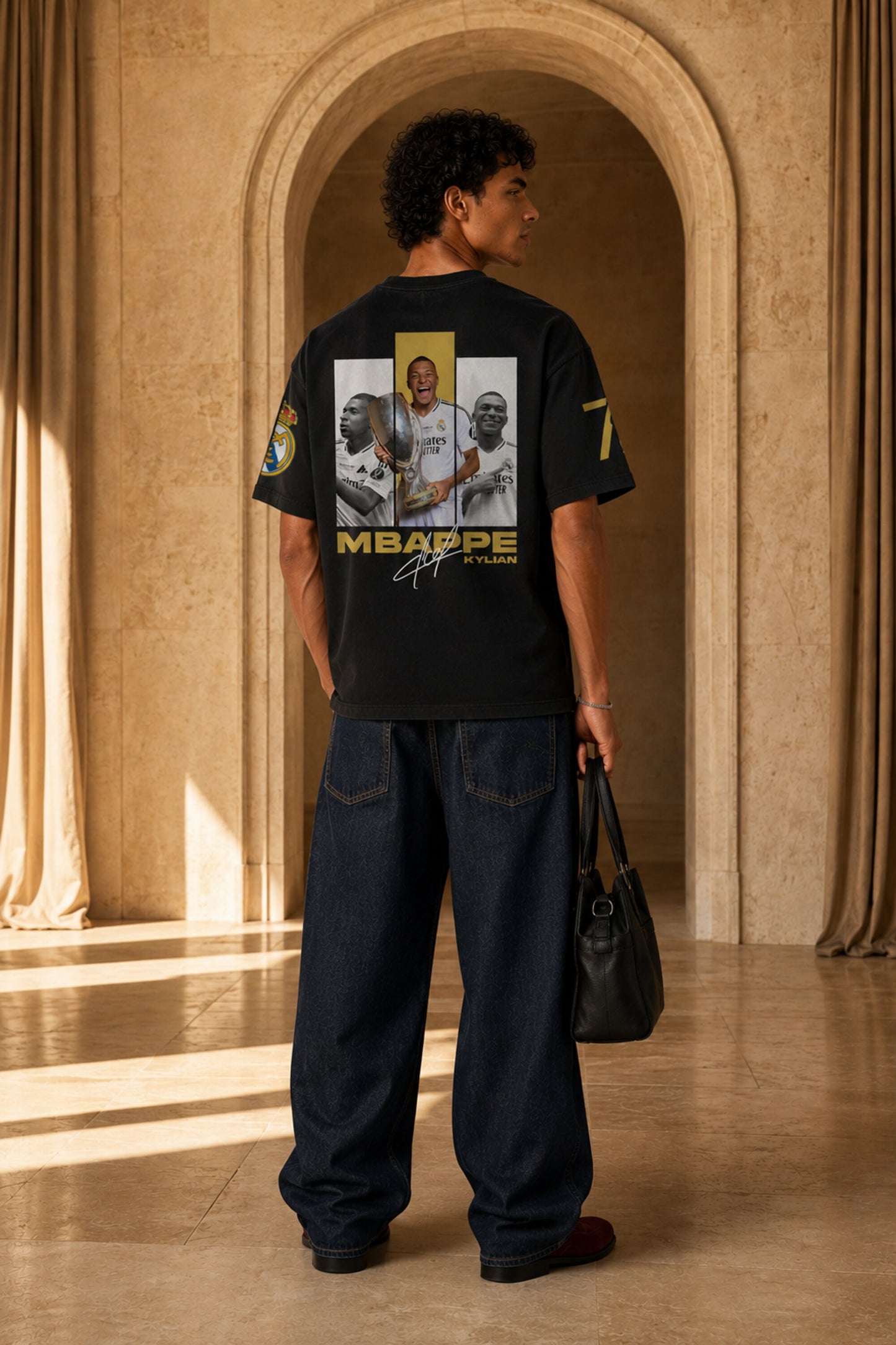 A man stands in a grand stone hall wearing a black graphic T-shirt from the back, which features a large photo collage and 'MBAPPE BY MANU' text, paired with loose dark blue jeans and holding a black bag.