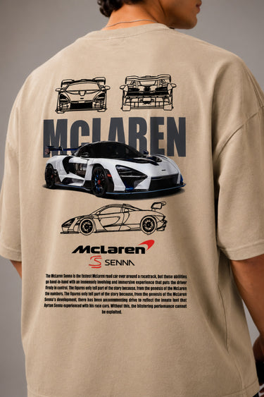 Man wearing a tan oversized t-shirt with a graphic print of the McLaren Senna car and "McLaren Senna" text. He is sitting on metal stairs.