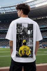 A man standing on a sports field in front of a stadium, wearing a white t-shirt with a large black-and-yellow graphic print on the back. The print features text reading 'FROM TICKET COLLECTOR TO TROPHY COLLECTOR' and a collage of cricket player images, including a prominent yellow jersey.