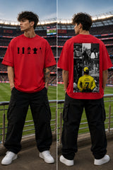 A man stands in a stadium wearing a red graphic T-shirt and black cargo pants, shown from both front and back. The front has a black trophy-style graphic across the chest, and the back features a large collage print with black photos and a yellow soccer player graphic.