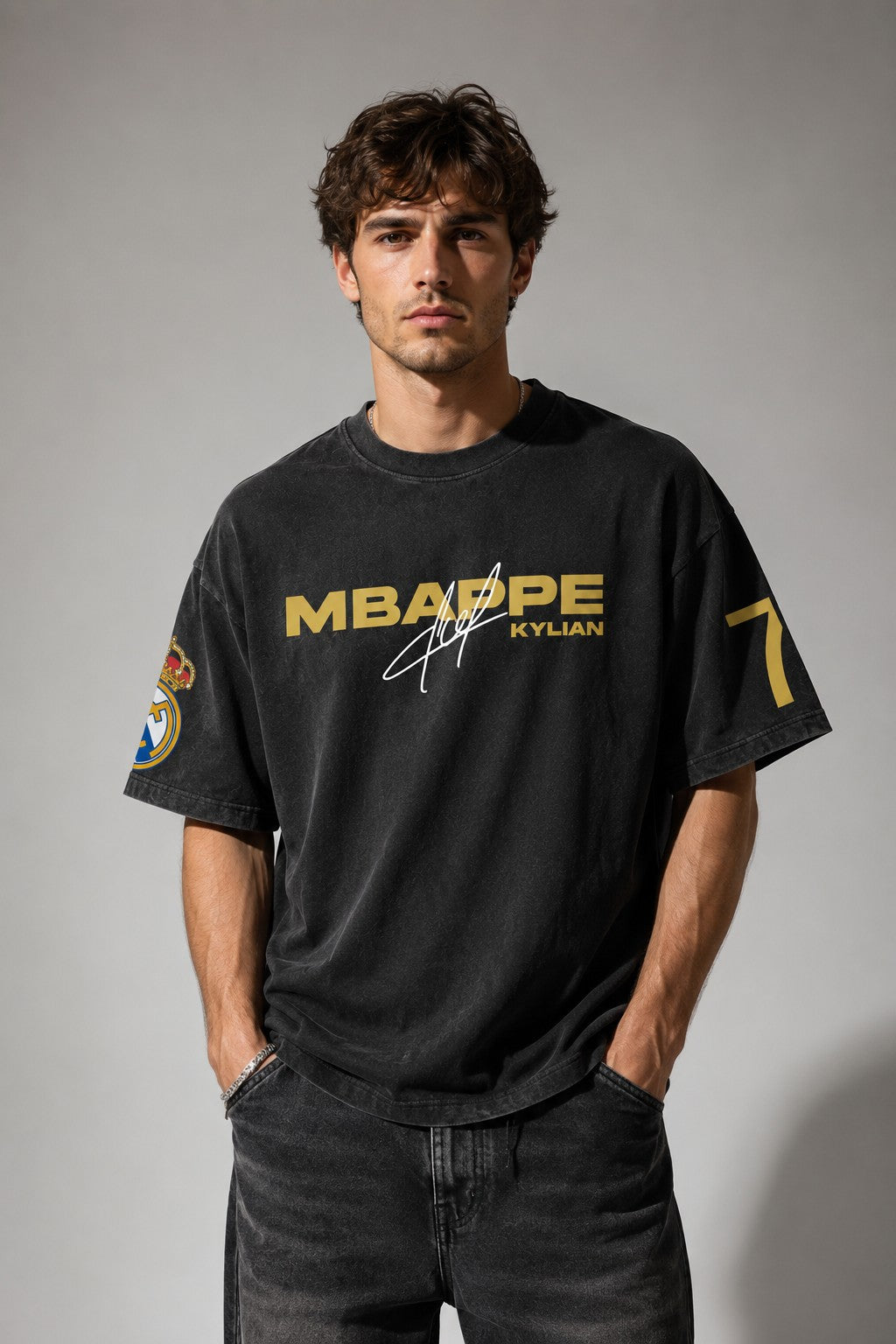 Model wearing an anthracite black Mbappe oversized t-shirt with gold "MBAPPE" graphic and signature, Real Madrid and number 7 logos on sleeves.