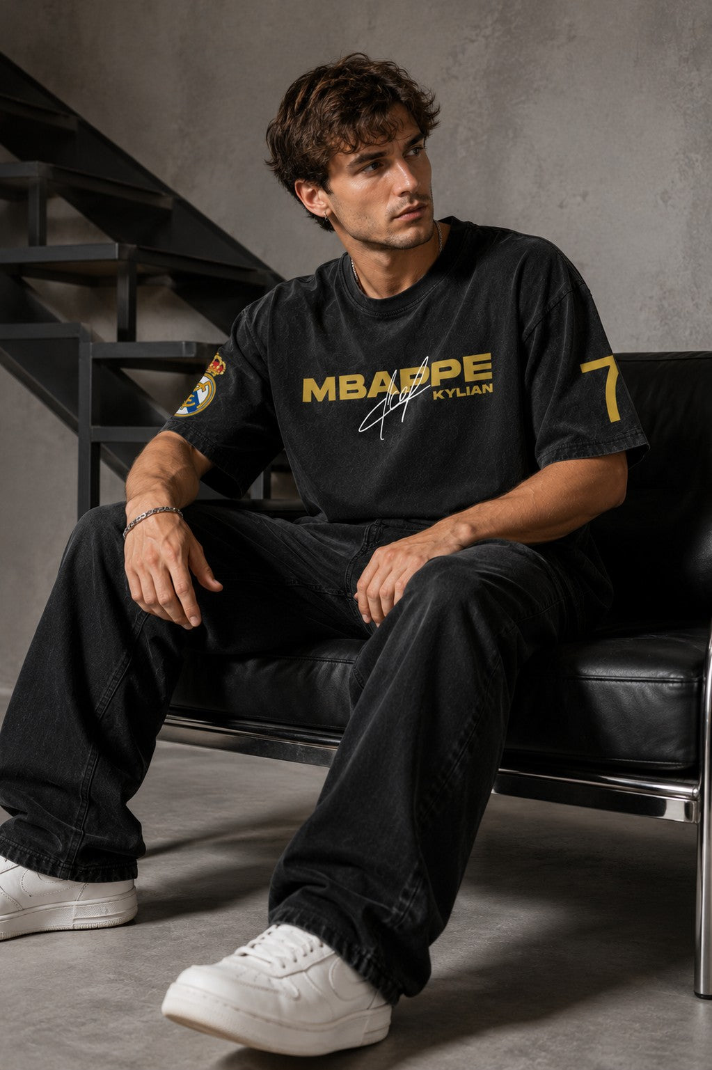 Model wearing an anthracite black Mbappe oversized t-shirt with gold "MBAPPE" graphic and signature, Real Madrid and number 7 logos on sleeves.