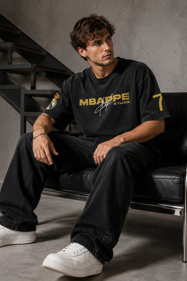 Model wearing an anthracite black Mbappe oversized t-shirt with gold "MBAPPE" graphic and signature, Real Madrid and number 7 logos on sleeves.