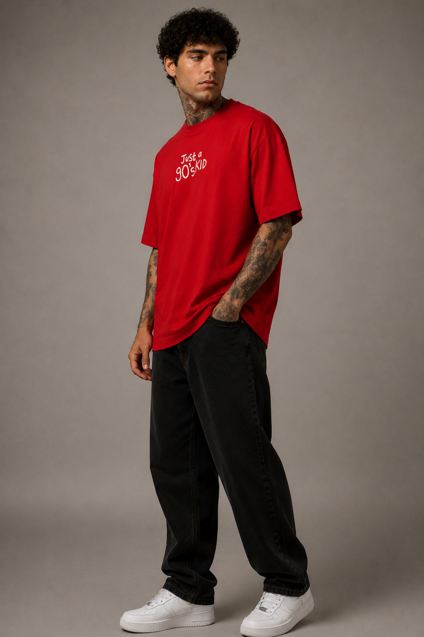 Model wearing a red oversized t-shirt with 'Just a 90's Kid' graphic. The tee has a relaxed fit, paired with black jeans and white sneakers, embodying a retro streetwear style.