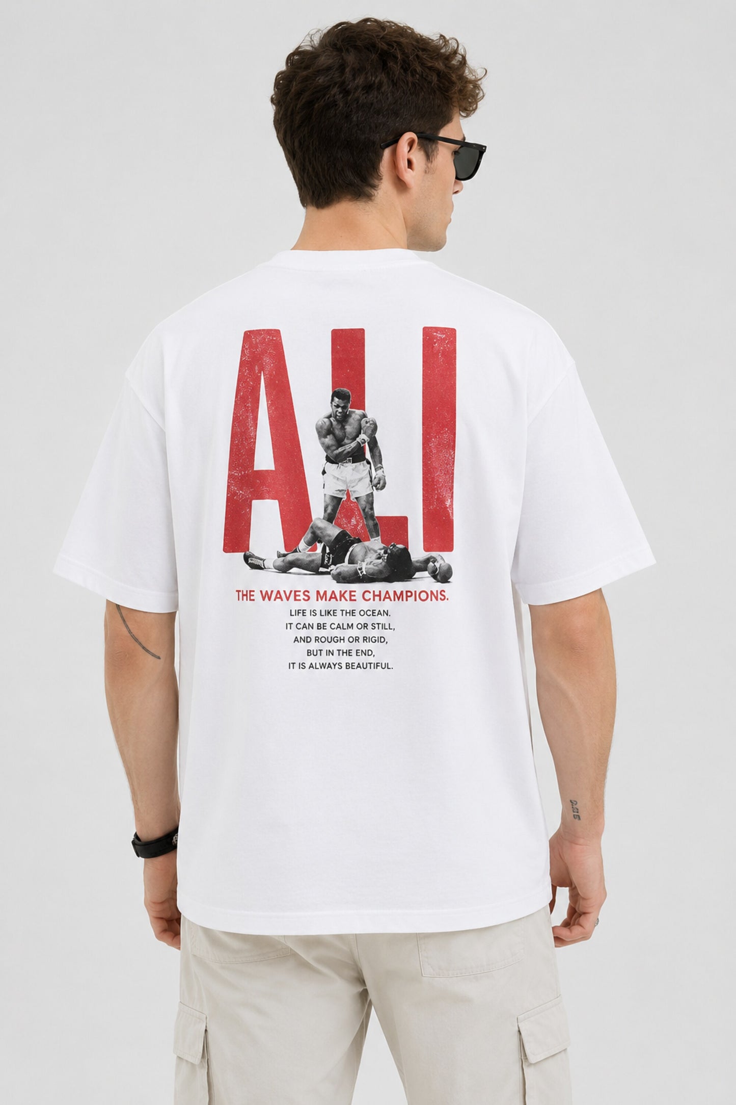 Man wearing a white ALI oversized t-shirt and beige cargo pants, sitting on grey stairs. He wears black sunglasses and jewelry. - white