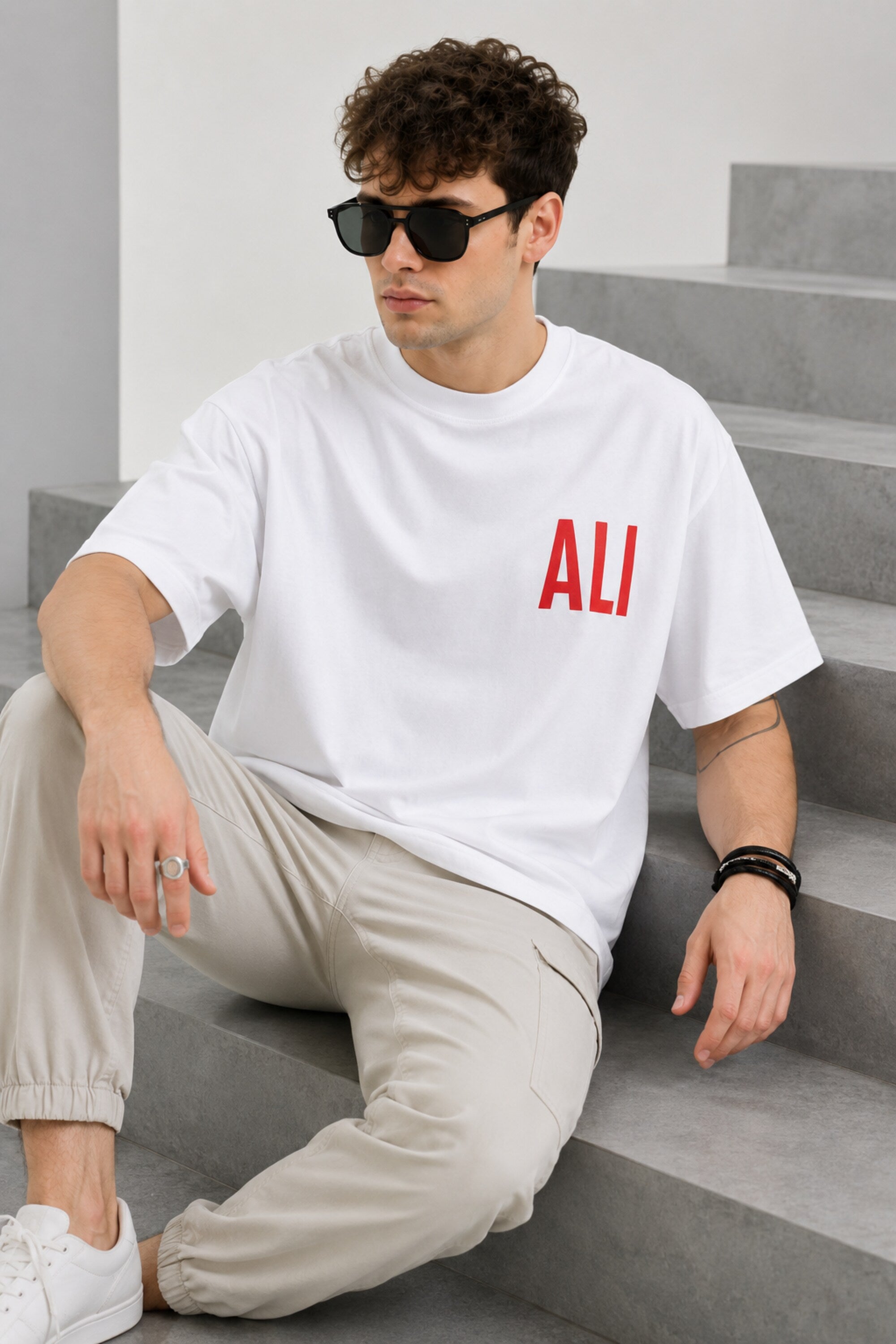Man wearing a white ALI oversized t-shirt and beige cargo pants, sitting on grey stairs. He wears black sunglasses and jewelry.