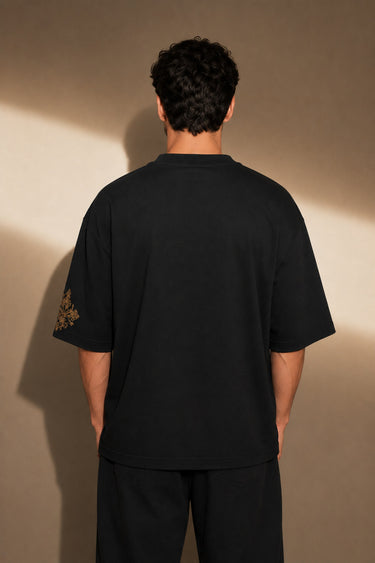 Back view of a person wearing a black ART ON OVERSIZED TSHIRT with gold embroidered detail on the left sleeve. Shows the oversized fit and premium streetwear aesthetic.