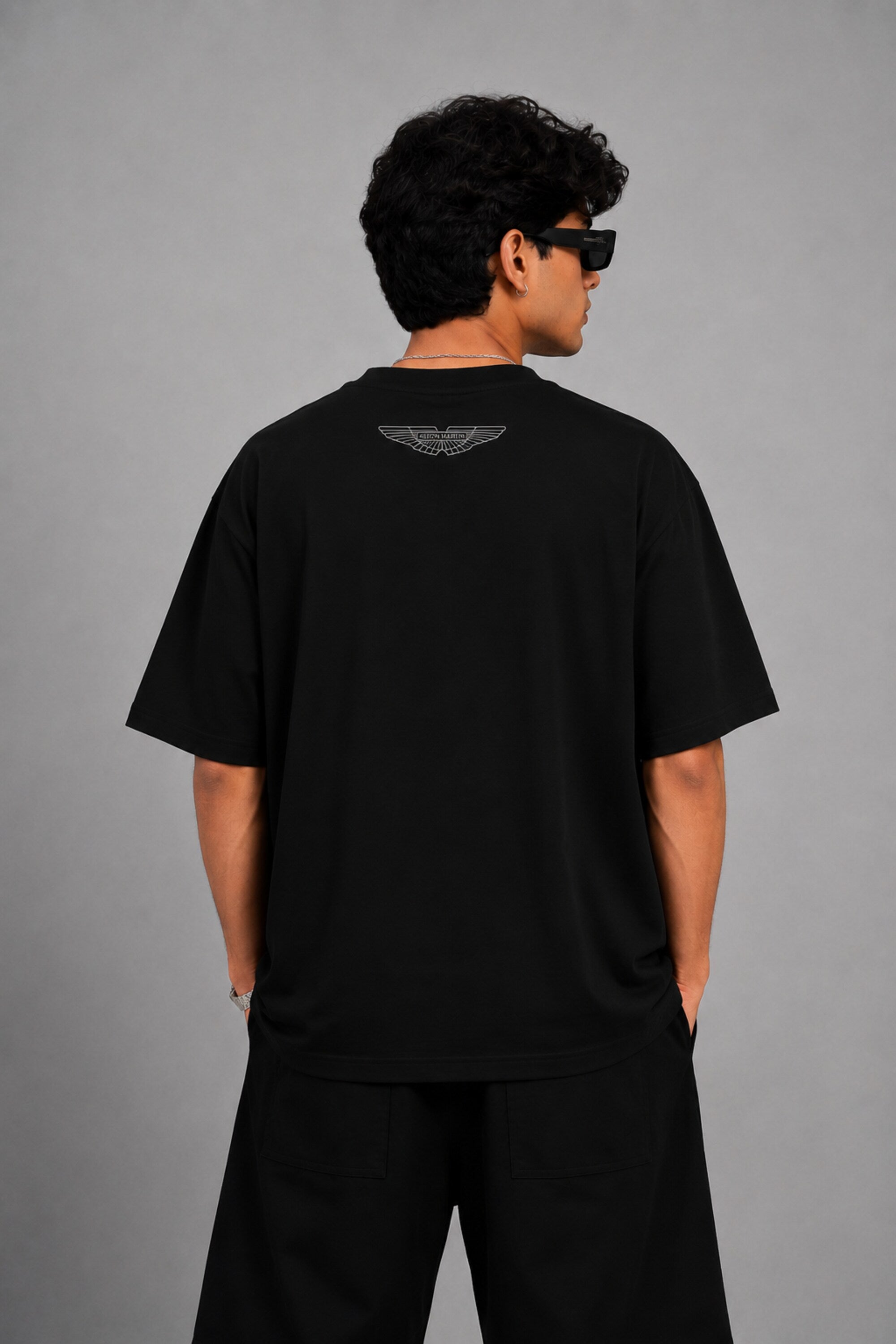 Man wearing a black Aston Martin oversized t-shirt with green racing stripes and the iconic Aston Martin wings logo, sitting on industrial stairs.