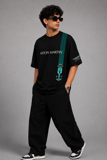 Man wearing a black Aston Martin oversized t-shirt with green racing stripes and the iconic Aston Martin wings logo, sitting on industrial stairs.
