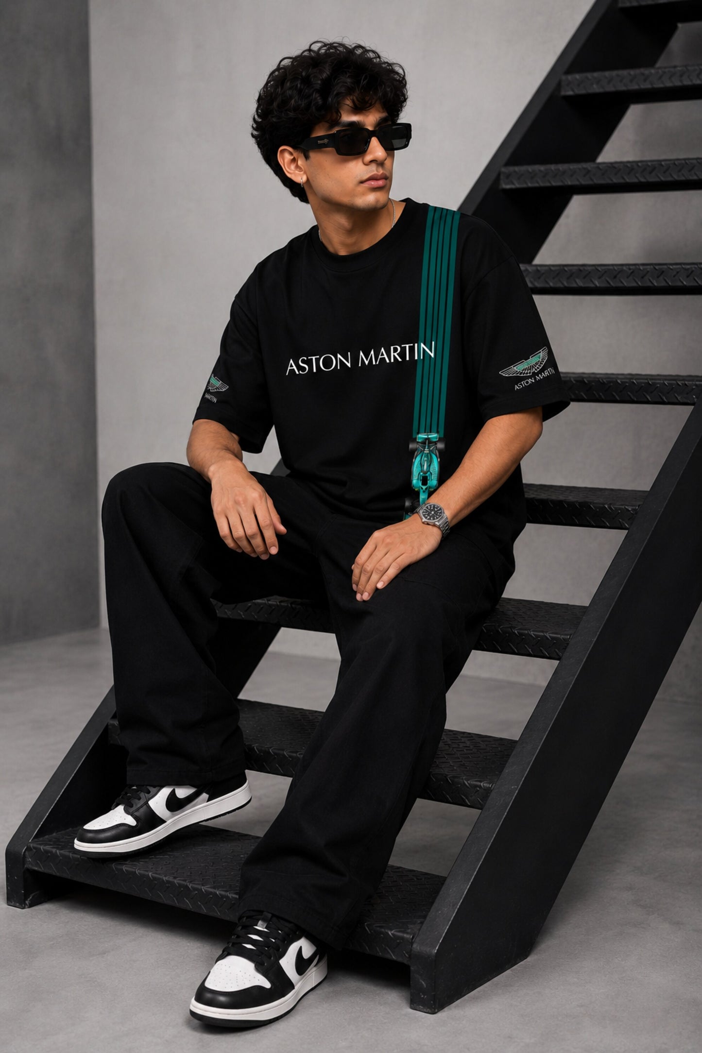 Man wearing a black Aston Martin oversized t-shirt with green racing stripes and the iconic Aston Martin wings logo, sitting on industrial stairs.