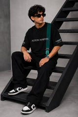 Man wearing a black Aston Martin oversized t-shirt with green racing stripes and the iconic Aston Martin wings logo, sitting on industrial stairs.