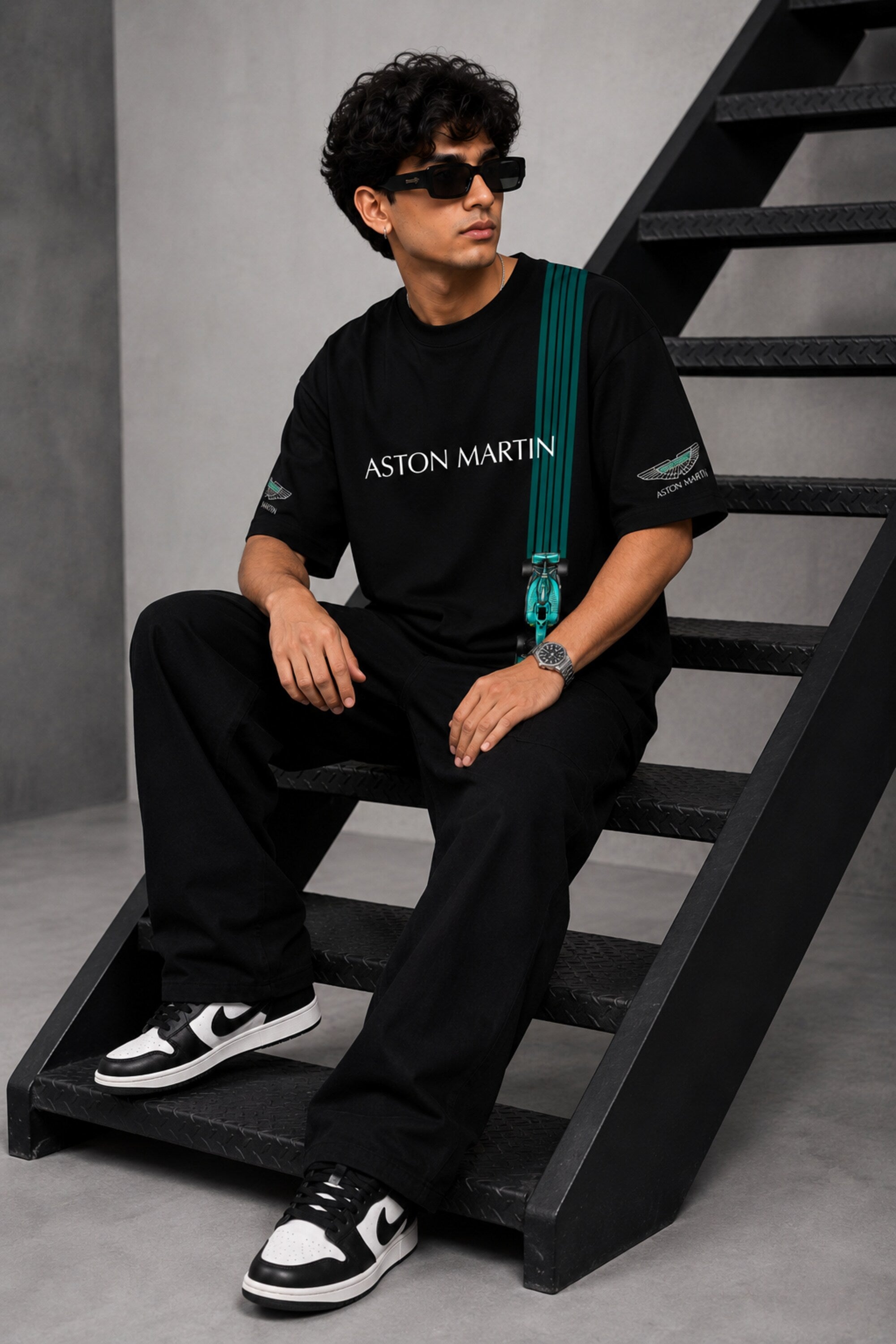 Man wearing a black Aston Martin oversized t-shirt with green racing stripes and the iconic Aston Martin wings logo, sitting on industrial stairs.