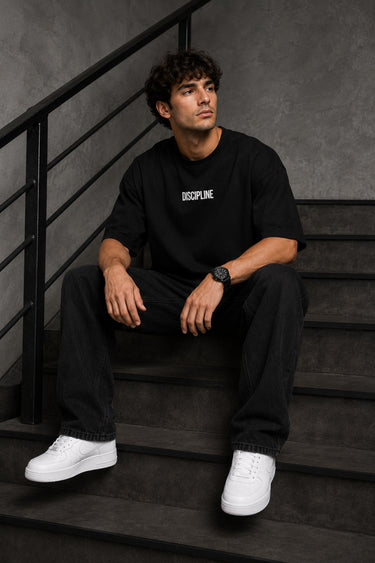 Man wearing a black DISCIPLINE WILD WOLF oversized t-shirt with white 'DISCIPLINE' text, dark wash jeans, and white sneakers, sitting on stairs.