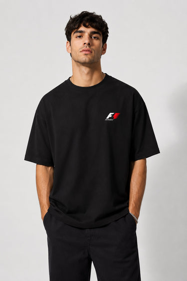 Model wearing a black oversized t-shirt with a small, embroidered Formula 1® logo in white, red, and gray on the chest. Casual streetwear style.
