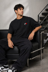 Model wearing a black oversized t-shirt with a small, embroidered Formula 1® logo in white, red, and gray on the chest. Casual streetwear style.
