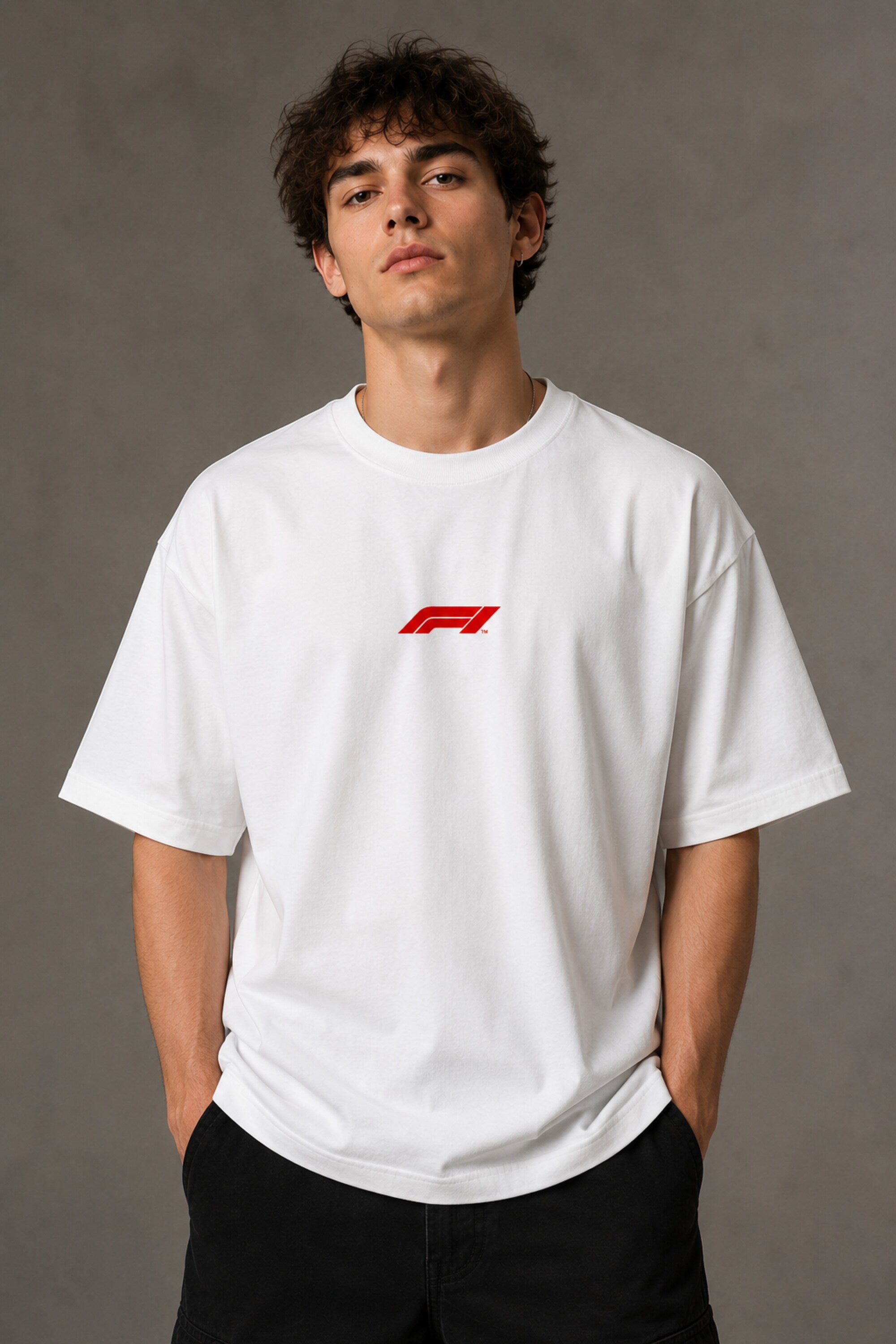 Man wearing white F1 Official oversized t-shirt with red F1 logo, black wide-leg jeans, and white sneakers, sitting on a black leather couch.