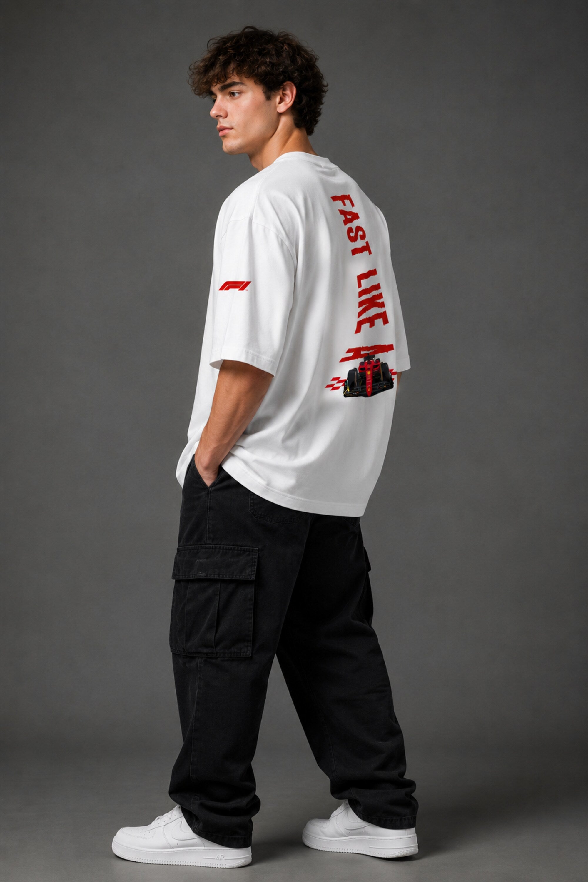 Man wearing white F1 Official oversized t-shirt with red F1 logo, black wide-leg jeans, and white sneakers, sitting on a black leather couch.