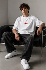 Man wearing white F1 Official oversized t-shirt with red F1 logo, black wide-leg jeans, and white sneakers, sitting on a black leather couch.