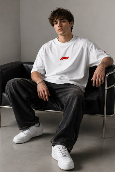 Man wearing white F1 Official oversized t-shirt with red F1 logo, black wide-leg jeans, and white sneakers, sitting on a black leather couch.