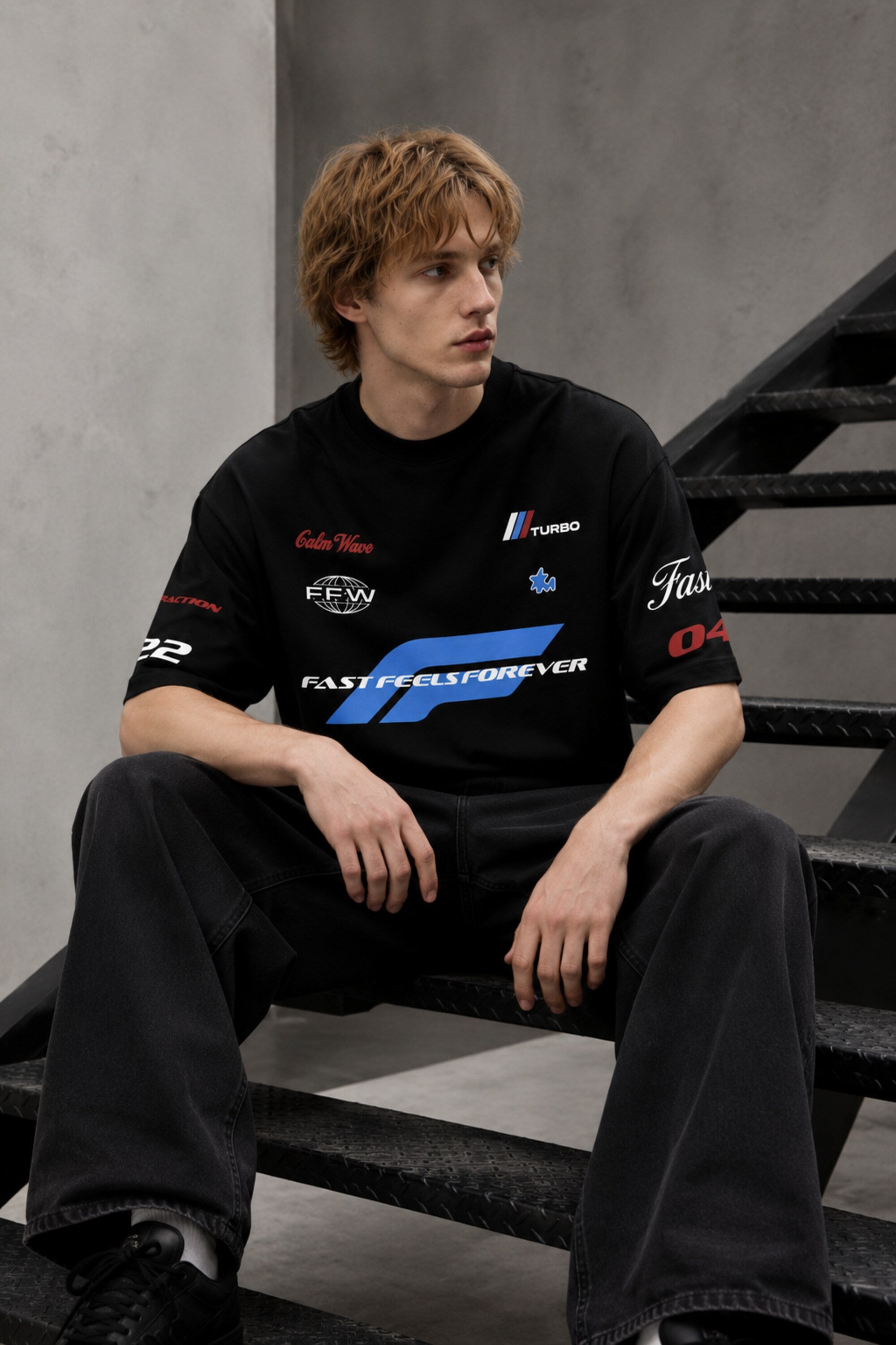 Model wearing a black Fast Feels Forever oversized t-shirt with graphic prints, sitting on a leather couch.