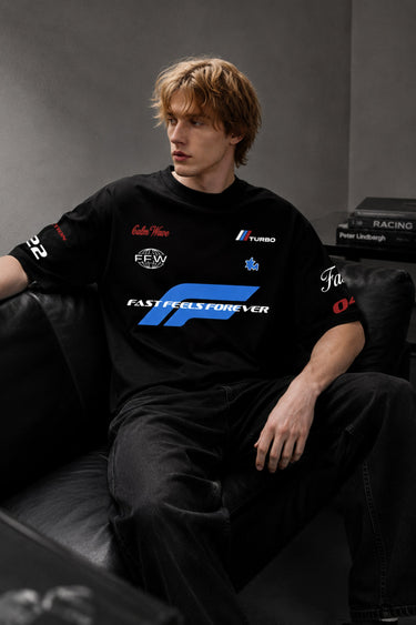 Model wearing a black Fast Feels Forever oversized t-shirt with graphic prints, sitting on a leather couch.
