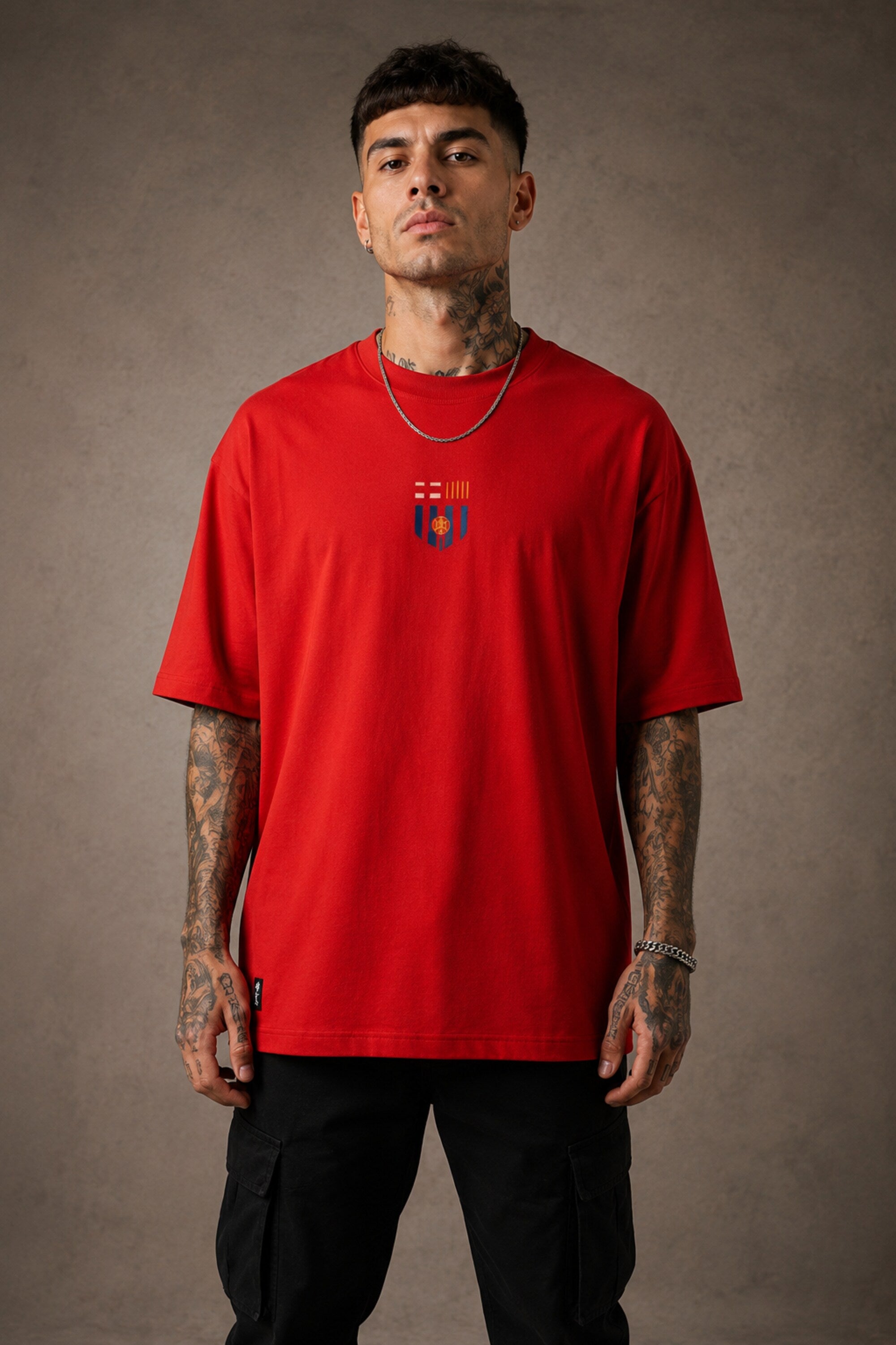 Man wearing a red FCB Messi fan oversized t-shirt, black pants, and white sneakers, sitting on a brown couch. - red