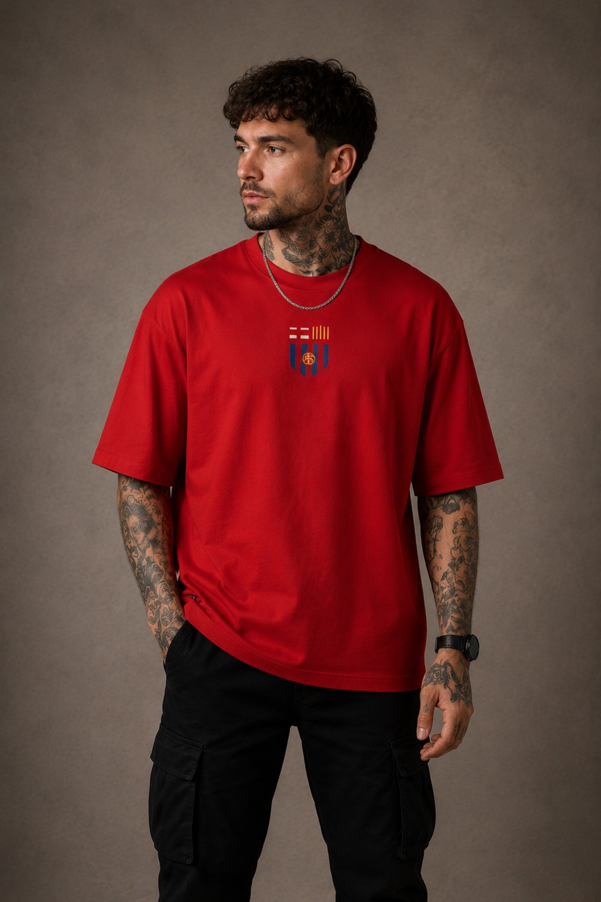 Man wearing a red FCB Messi fan oversized t-shirt, black pants, and white sneakers, sitting on a brown couch.