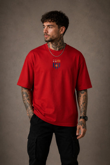 Man wearing a red FCB Messi fan oversized t-shirt, black pants, and white sneakers, sitting on a brown couch.