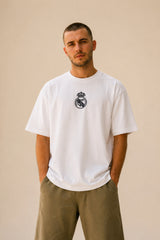 Man wearing a white oversized Real Madrid t-shirt with a circular club crest logo, paired with olive green pants and black sneakers, posing casually with hands in pockets. - white