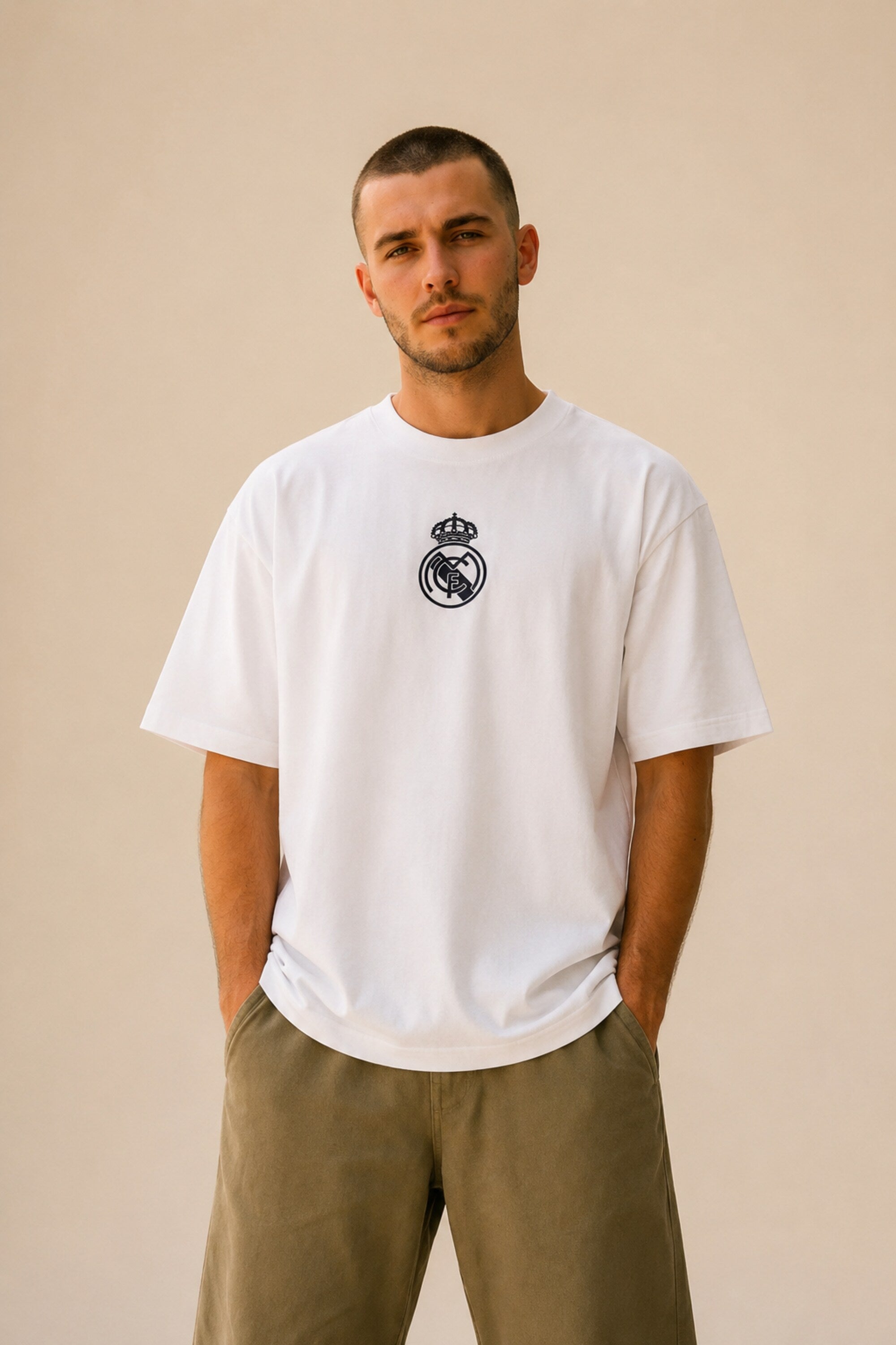 Man wearing a white oversized Real Madrid t-shirt with a circular club crest logo, paired with olive green pants and black sneakers, posing casually with hands in pockets. - white