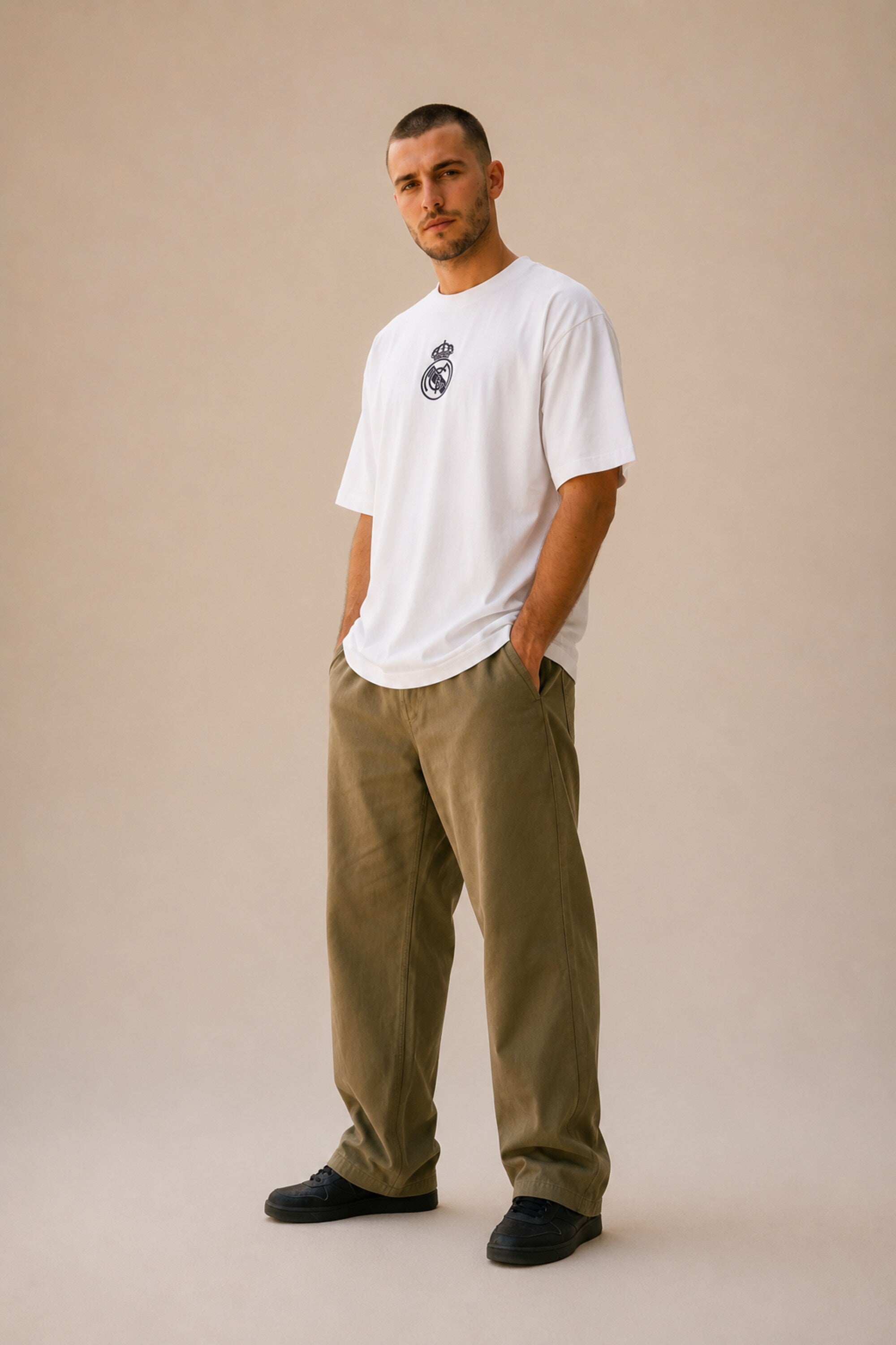 Man wearing a white oversized Real Madrid t-shirt with a circular club crest logo, paired with olive green pants and black sneakers, posing casually with hands in pockets.