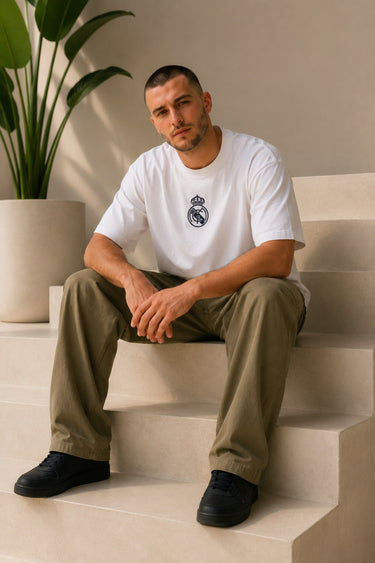 Man wearing a white oversized Real Madrid t-shirt with a circular club crest logo, paired with olive green pants and black sneakers, posing casually with hands in pockets.