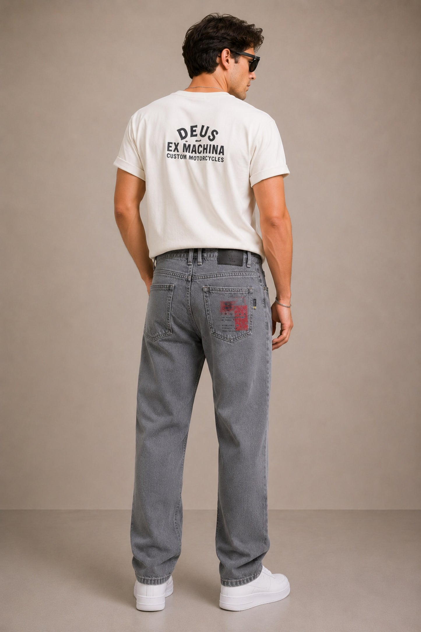 Man wearing a white t-shirt with 'Deus Ex Machina' logo and gray jeans on a beige background