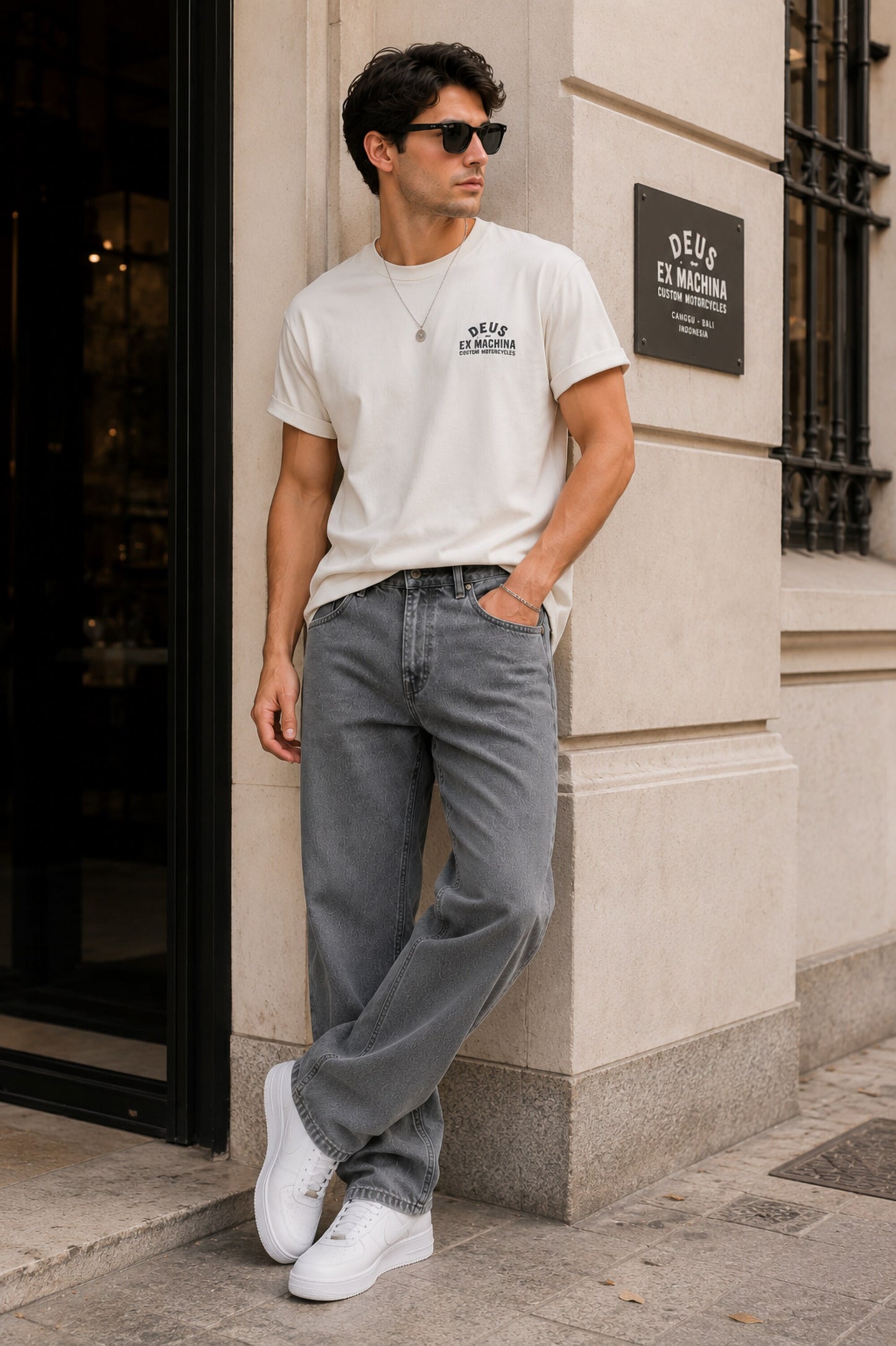 Man wearing a white t-shirt and gray jeans standing against a building wall.