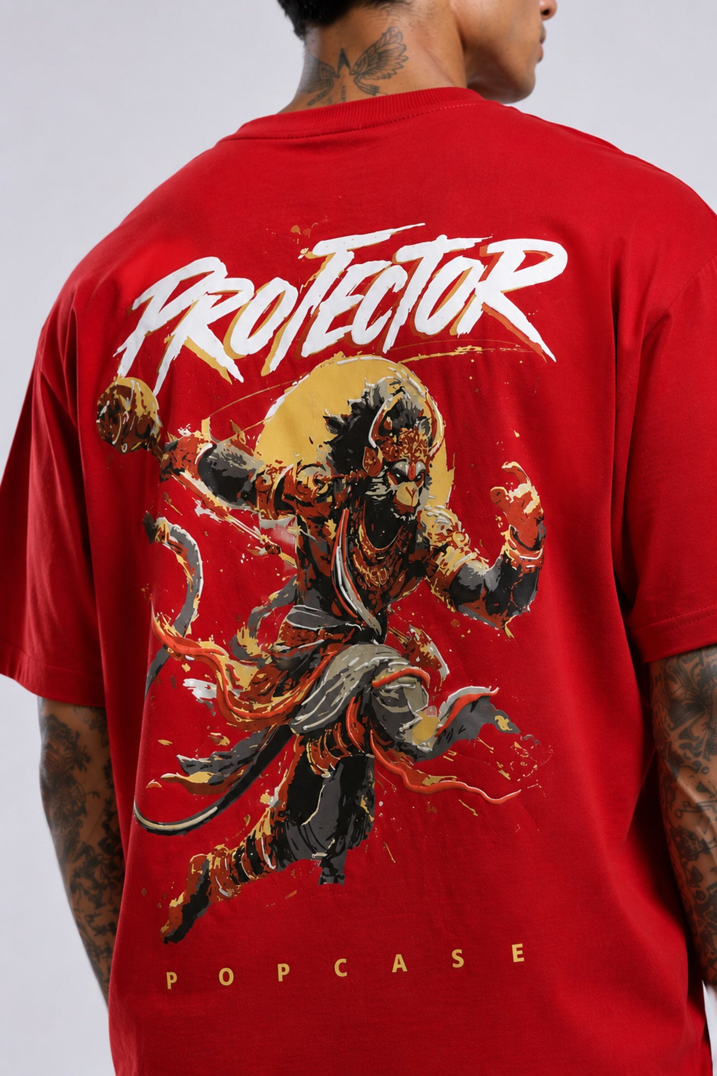 Red oversized t-shirt with 'PROJECTOR HANUMANJI' graphic design of a warrior monkey god and 'POPCASE' logo, worn by a man.