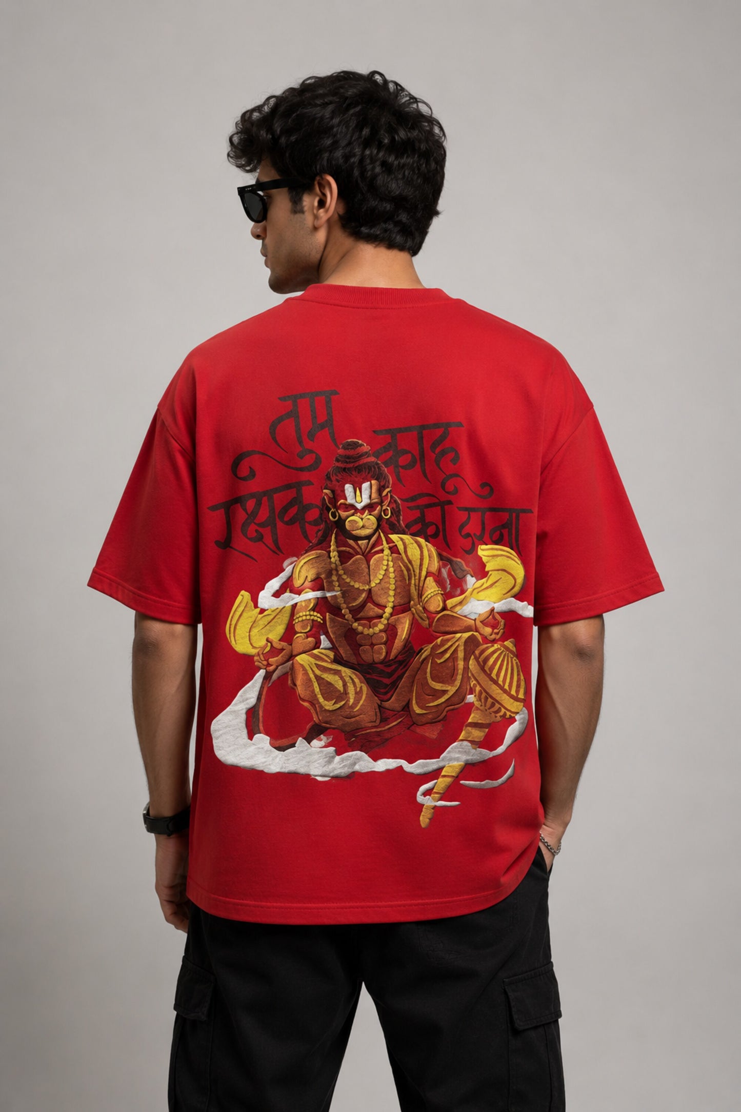 Man wearing a red PROJECTOR HANUMANJI oversized t-shirt with black pants and white sneakers, sitting on metal stairs.