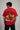 Man wearing a red PROJECTOR HANUMANJI oversized t-shirt with black pants and white sneakers, sitting on metal stairs.