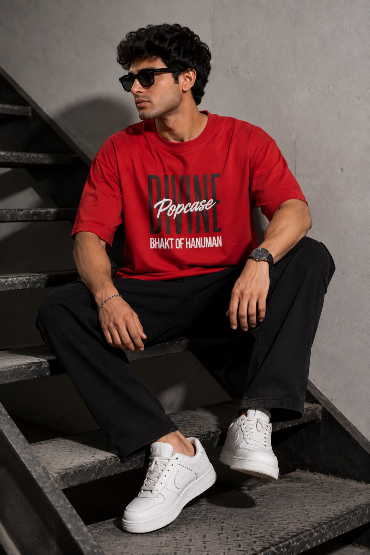 Man wearing a red PROJECTOR HANUMANJI oversized t-shirt with black pants and white sneakers, sitting on metal stairs.