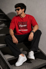 Man wearing a red PROJECTOR HANUMANJI oversized t-shirt with black pants and white sneakers, sitting on metal stairs.