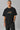 Back view of a man wearing a black HORN OK PLEASE oversized t-shirt with gold 'POPCASE' logo on the upper back.
