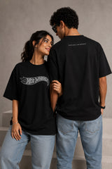 A couple wearing matching black oversized Hot Wheels t-shirts and light wash jeans, posing intimately against a neutral background.