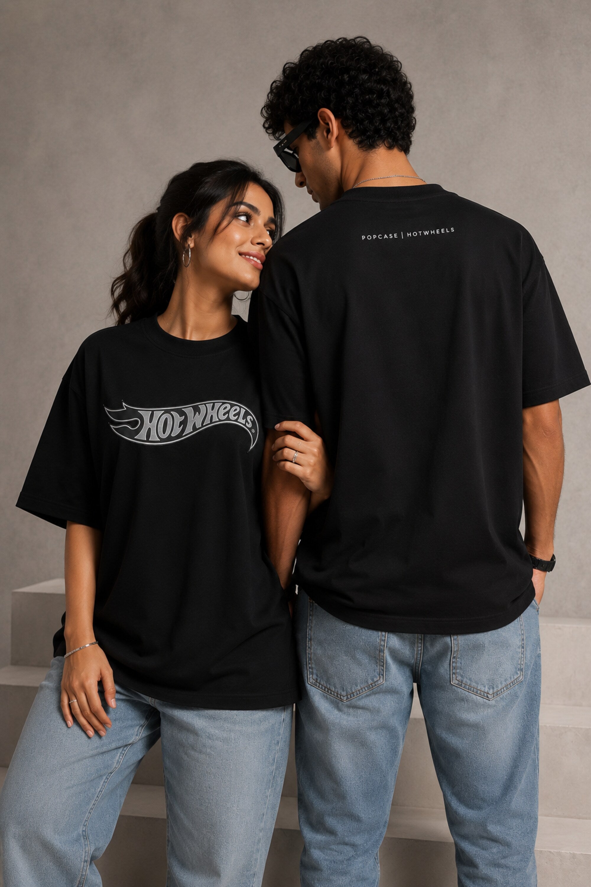 A couple wearing matching black oversized Hot Wheels t-shirts and light wash jeans, posing intimately against a neutral background.