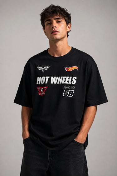 Man wearing a black STAR BOY oversized t-shirt with "Hot Wheels" graphics, sitting on a black couch. Streetwear fashion. - black