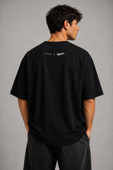 Man wearing a black STAR BOY oversized t-shirt with "Hot Wheels" graphics, sitting on a black couch. Streetwear fashion.