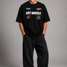Man wearing a black STAR BOY oversized t-shirt with "Hot Wheels" graphics, sitting on a black couch. Streetwear fashion.