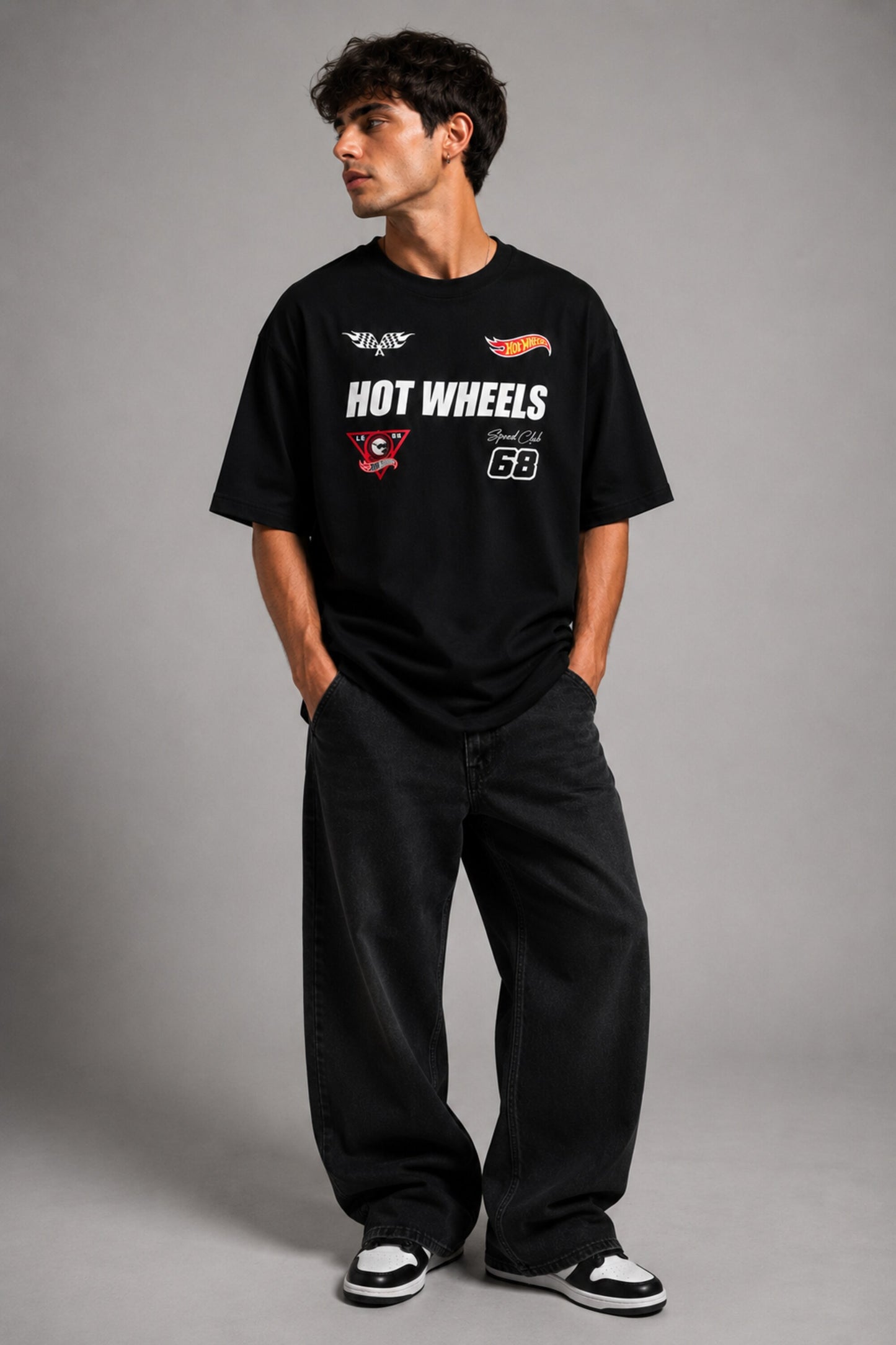 Man wearing a black STAR BOY oversized t-shirt with "Hot Wheels" graphics, sitting on a black couch. Streetwear fashion.