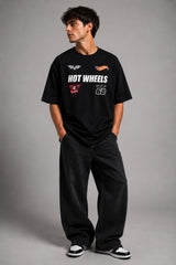 Man wearing a black STAR BOY oversized t-shirt with "Hot Wheels" graphics, sitting on a black couch. Streetwear fashion.