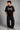 Man wearing a black STAR BOY oversized t-shirt with "Hot Wheels" graphics, sitting on a black couch. Streetwear fashion.